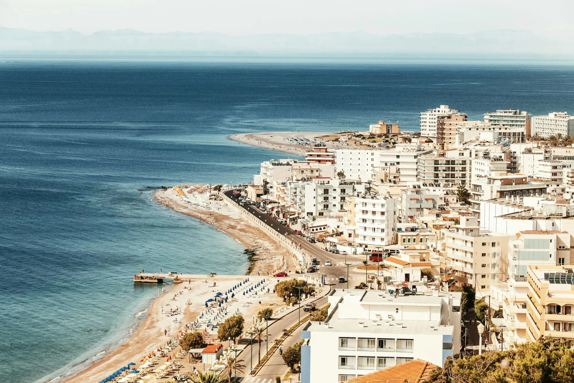 Aerial view of a coastal city with white buildings and a beachside promenade along the blue sea.