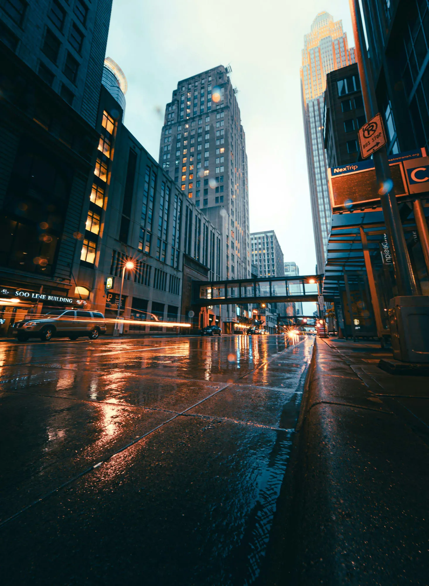 A rainy downtown street with tall buildings and reflective streetlights creating a moody atmosphere.
